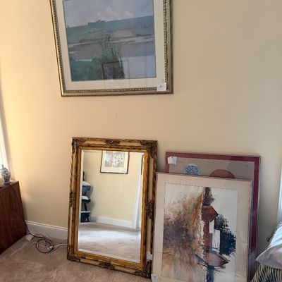 Large ornate gold-framed wall mirror leaning against a wall next to framed watercolor landscape artwork, with a framed landscape print...