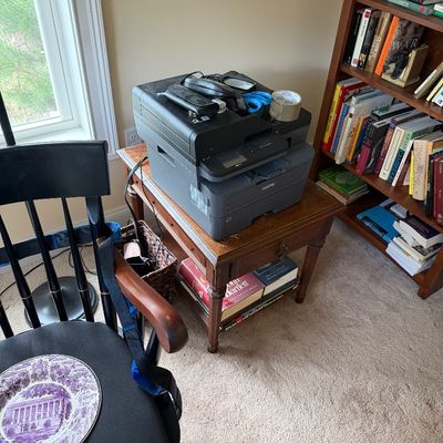 Brother all-in-one laser printer sitting on a vintage wooden end table with a lower shelf, shown alongside a black wooden armchair.