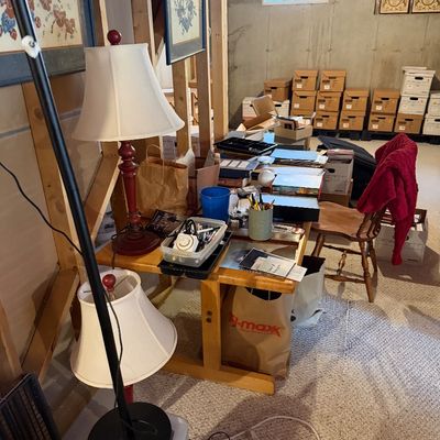 Wooden trestle desk paired with a spindle-back chair, featuring various office supplies, table lamps, and framed artwork in a basement...