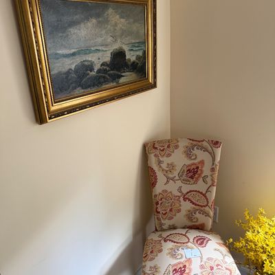 Framed oil painting of a rocky seascape in an ornate gold frame, paired with a high-back accent chair featuring a red and beige floral...