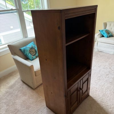 Tall wooden media cabinet or bookcase featuring open upper shelving and a lower two-door storage compartment, shown in a living room...