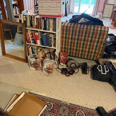 Assorted household items including a white wooden bookshelf filled with books, a plaid upholstered armchair, power tools, a wood-framed...