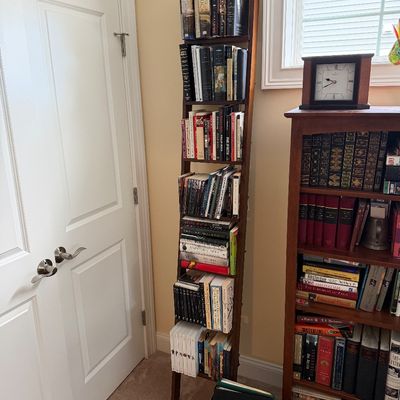 Tall, narrow wooden ladder-style bookshelf filled with books, standing next to a traditional wooden bookcase.