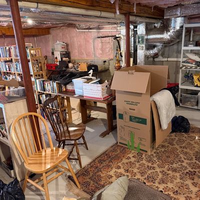 Unfinished basement storage area featuring wooden Windsor-style chairs, a patterned area rug, various storage boxes, and bookshelves...