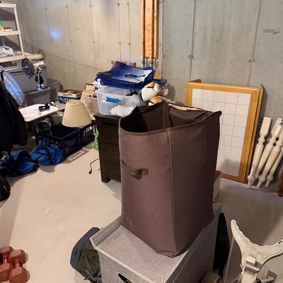 Assorted household items in a basement, including a brown fabric laundry hamper, a grey storage bin, a golf push cart, and various...