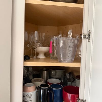 Assorted kitchenware including ceramic mugs, wine glasses, a plastic measuring cup, and mixing bowls arranged inside a wooden cabinet.