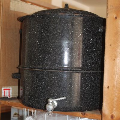 Vintage black speckle enamelware stock pot or spigoted water dispenser sitting on a wooden shelf, showing minor light wear.
