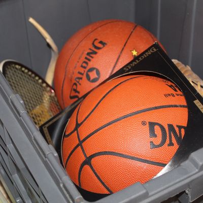 Plastic storage crate containing vintage sports equipment, including two Spalding basketballs, a tennis racket, and a baseball glove.