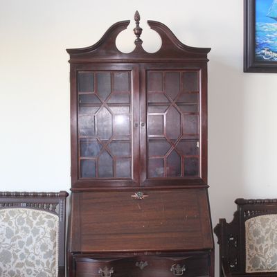 Traditional dark wood slant-front secretary desk featuring a pediment top with central finial, glass-front upper cabinet, and serpentine...