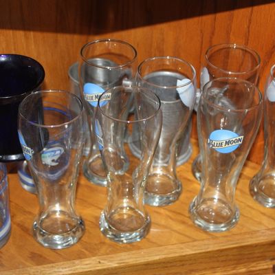 Collection of branded glassware displayed in a wooden cabinet, including Blue Moon pilsner glasses, Seattle Seahawks glasses, pewter...
