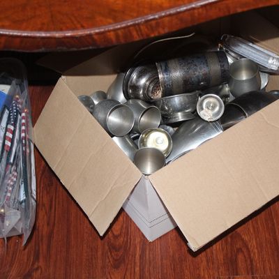 Cardboard box filled with vintage pewter and metal barware items, including goblets, shakers, and jiggers.