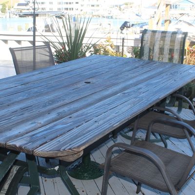 Rustic weathered wood plank patio dining table with umbrella hole, set on a metal base alongside surrounding outdoor mesh armchairs.