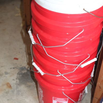 Stack of 5-gallon plastic utility buckets, including several red Harbor Freight buckets and one white bucket, with metal wire handles.