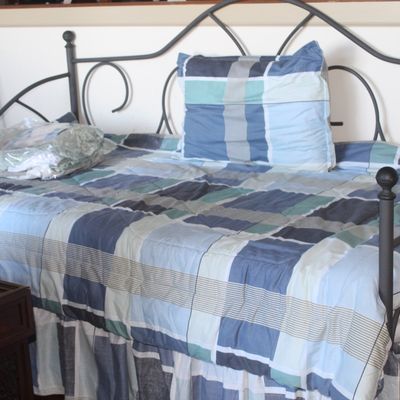 Black metal scrollwork daybed with finial accents, dressed in a blue and teal plaid bedding ensemble including a matching bedskirt and...
