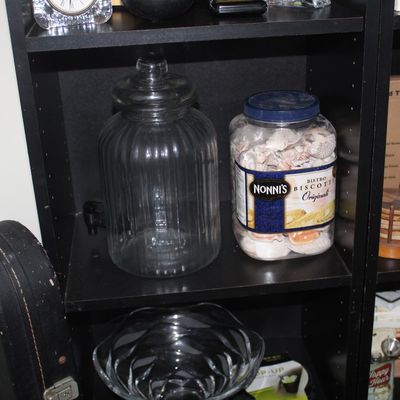 Black wooden shelving unit holding a large ribbed clear glass jar with lid, a Nonni's biscotti tub repurposed for seashell storage,...