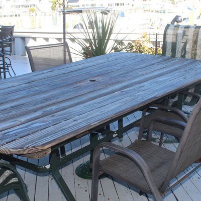 Rustic weathered wood-top patio dining table with an umbrella hole, accompanied by several metal mesh outdoor chairs on a deck.