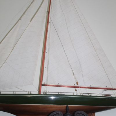 Detailed wooden sailboat model with a green painted hull, wooden base, and full cloth sails, displayed atop a piece of furniture.