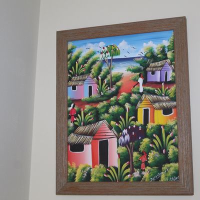 Vibrant Caribbean naive art oil painting depicting a coastal village with colorful huts, housed in a textured wood frame.