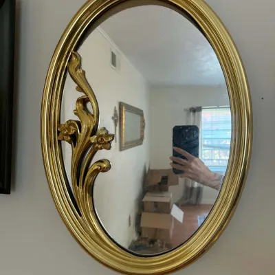 Vintage Syroco-style oval wall mirror featuring a bright gold-finished frame with an elegant floral and scrollwork overlay on the left side.