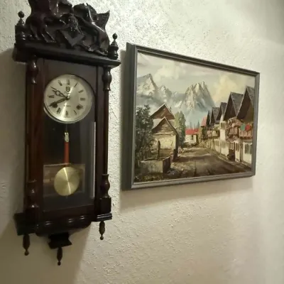 Vintage wooden regulator wall clock with ornate horse carvings on top, alongside a framed oil painting depicting an alpine village street...
