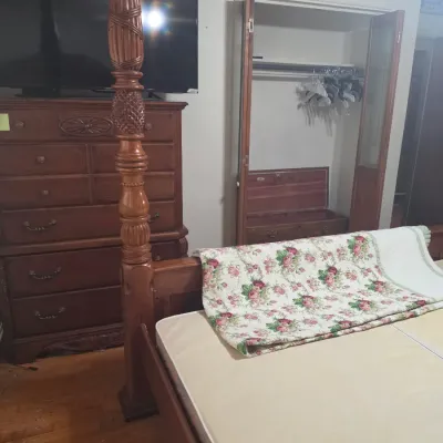 Traditional four-poster wooden bed frame with carved pineapple details, shown alongside a matching tall chest of drawers and a floral quilt.