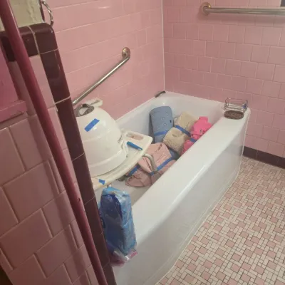 Vintage mid-century bathroom featuring pink wall tile, mosaic floor tile, and a white bathtub filled with various household items,...