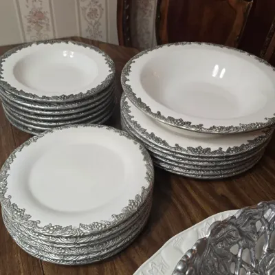 Stacks of white ceramic dinnerware featuring ornate, embossed silver-tone metal rims with a grape and vine motif.