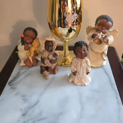 Four small Black angel and child figurines arranged on a marble-top table in front of a tall, gold-plated chalice.