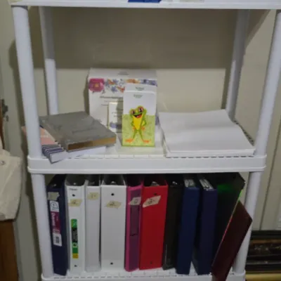Four-tier white plastic utility shelving unit holding office supplies, binders, and small electronics.