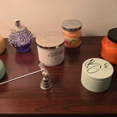 Collection of scented candles in various jars and tins, paired with a silver-tone candle snuffer and a decorative purple glass perfume...