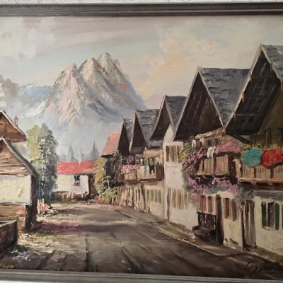 Framed oil painting depicting a European alpine village street with mountains in the background, signed by the artist in the lower right...