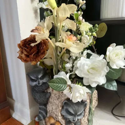 Whimsical ceramic tree trunk planter featuring two sculpted raccoons, filled with an assortment of faux white and brown silk flowers.