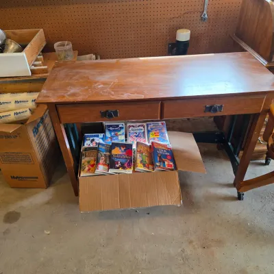 Sale Photo Thumbnail #69: Wooden writing desk with two drawers and metal pulls, shown with a cardboard box of vintage Disney VHS tapes underneath.