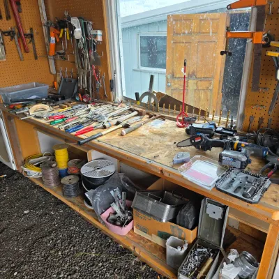 Sale Photo Thumbnail #25: Wooden workbench cluttered with various hand tools, power tools, hardware, and spools of wire, set against a pegboard wall in a workshop.