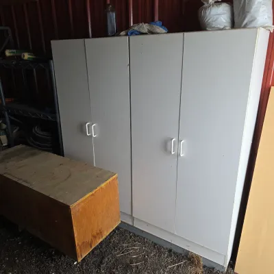 Sale Photo Thumbnail #90: Two white laminate double-door storage cabinets with white plastic handles, shown alongside a wooden storage box in a garage or shed setting.