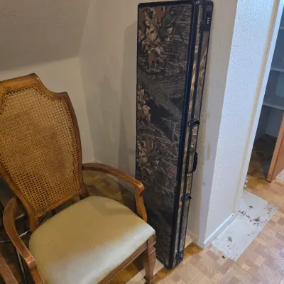 Sale Photo Thumbnail #11: Hard-sided rifle or shotgun case with a camouflage pattern, standing next to a vintage wooden armchair with a cane back and upholstered seat.