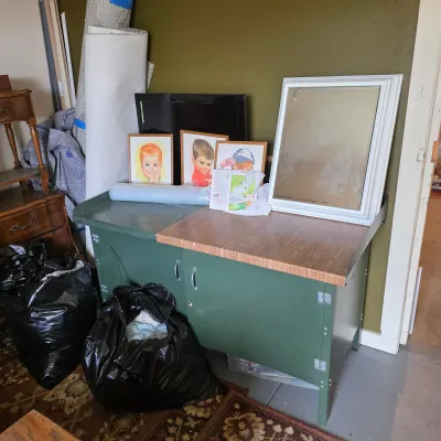 Sale Photo Thumbnail #5: Vintage green metal workbench with a wood-look laminate top, holding a white-framed mirror, framed children's portraits, and other miscellaneous items.