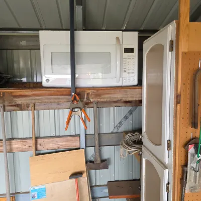 Sale Photo Thumbnail #26: White GE over-the-range microwave oven stored on a wooden shelf in a shed, alongside various tools and hardware.