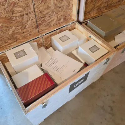 Sale Photo Thumbnail #58: Large wooden shipping crate filled with boxed items, including white gift boxes with silver labels and a red striped box, accompanied by an inventory list.