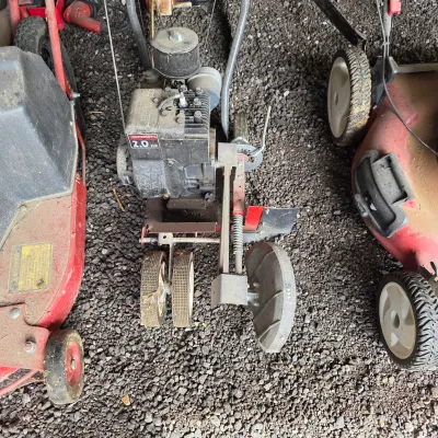 Sale Photo Thumbnail #54: Vintage Mastercut 2.0 HP gas-powered lawn edger, showing signs of use and wear, flanked by two red lawn mowers on a gravel surface.