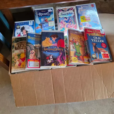 Sale Photo Thumbnail #70: Collection of vintage Disney VHS tapes in clamshell cases, including titles like Sleeping Beauty, Robin Hood, and Old Yeller, stored in a cardboard box.