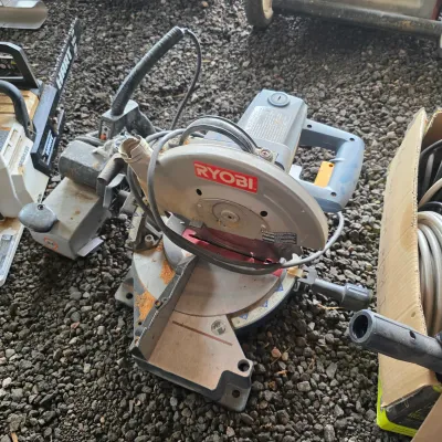Sale Photo Thumbnail #44: Ryobi compound miter saw showing some sawdust and wear from use, displayed on a gravel surface alongside other outdoor power tools.