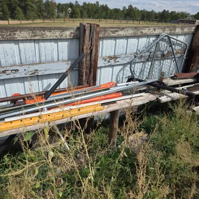 Sale Photo Thumbnail #81: Assorted outdoor farm and fencing equipment, including metal posts, a gate, and orange tools, resting on a wooden bench against a fence.