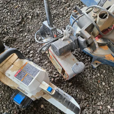 Sale Photo Thumbnail #45: Assorted power tools including a brushless chainsaw, a vintage Black & Decker belt sander, and a miter saw, showing signs of heavy use and sawdust.