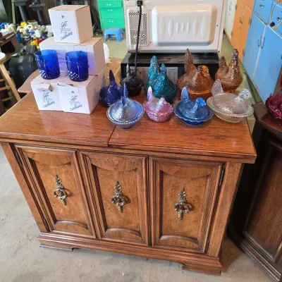 Sale Photo Thumbnail #73: Vintage wooden credenza with ornate metal pulls, displaying a collection of colorful glass hen-on-nest dishes and boxed blue glassware.