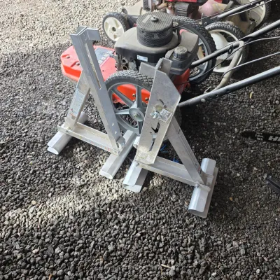 Sale Photo Thumbnail #50: Pair of aluminum scaffolding trestles or ladder jacks, shown alongside a walk-behind lawn mower on a gravel surface.