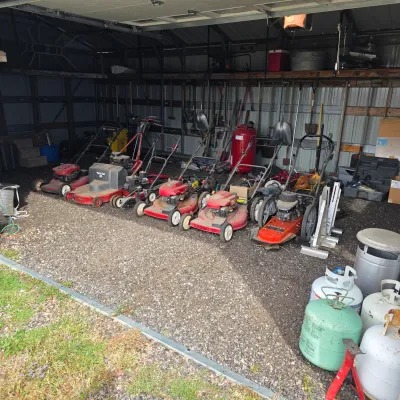 Sale Photo Thumbnail #23: Collection of vintage and modern walk-behind lawn mowers, including Toro and Snapper models, displayed alongside yard tools and propane tanks in a garage.