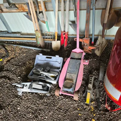 Sale Photo Thumbnail #31: Assorted garage tools including a pink hydraulic floor jack, a smaller grey floor jack in its case, a scissor jack, and various long-handled garden tools leaning against a wall.