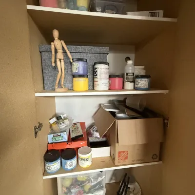 various art supplies, including acrylic paint jars, a mannequin, plastic storage bins, and brushes.