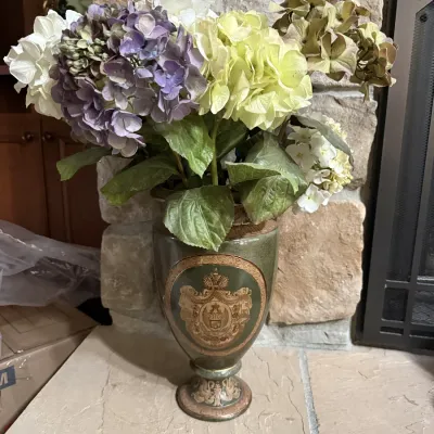 Ornate ceramic urn-style vase featuring a gold-toned heraldic crest motif, filled with colorful artificial hydrangea blossoms.